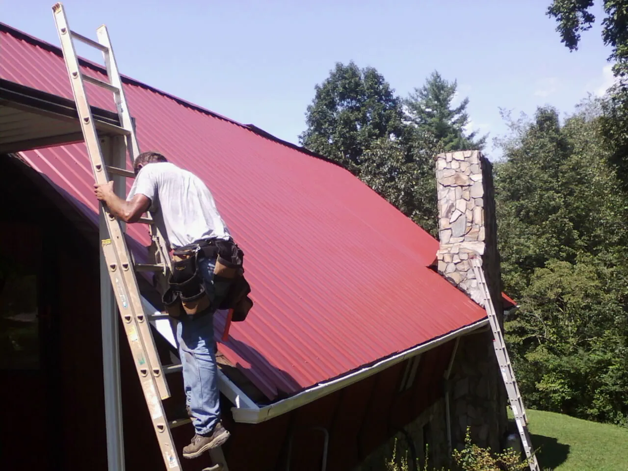 Roofer on a ladder beside a red metal roof and stone chimney — Soffit & Fascia Repair by Roofing Brothers in Mulberry